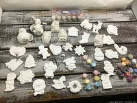 Photo showing a variety of unpainted white ceramic figurines arranged on a table with small paint pots and brushes above and to the right