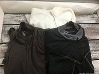Photo of three ladies sweaters folded and laid side by side: white, dark brown, and black with grey collar.