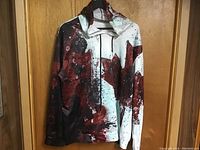 Front view of maple leaf design polyester hoodie hanging on door. Large red maple leaf centered, multi-colored splatter background.