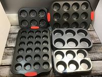 Five metal muffin and cupcake pans arranged on a wooden surface, showing the variety of sizes and some with red silicone grip handles.