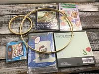 Two wooden embroidery hoops and multiple unopened embroidery and needlepoint kits with various designs displayed on rustic wooden surface.