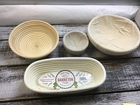 Four proofing dough baskets of various sizes placed on wooden background. Includes oval Banneton basket with label, two round rattan baskets, and a smaller fabric-lined basket. Showcases baskets from top and angled views to highlight size and spiral detail.