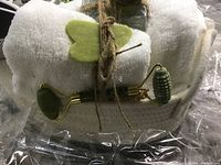 Close-up of cotton rope basket with white towel, green jade facial roller and gua sha stone tied with jute twine, inside clear cellophane wrapping.