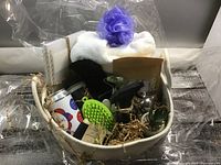 Overview of spa basket with visible towel, bath pouf, black pouch, can, green massager and other contents wrapped in cellophane