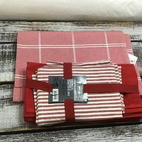 Table linens pictured on wooden surface consisting of pink checkered table runner, red placemats, and boxed set of 8 red and white striped tea towels tied with red ribbon.
