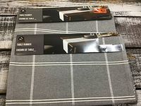 Photo showing both table runners folded with packaging labels on top, gray fabric with white checkered lines visible.