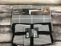Grey table runner with a white grid pattern, folded with packaging label showing size and product info. Pack of black and white striped placemat and napkin set also visible.