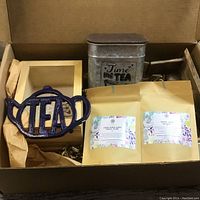 Box with tea tin labeled 'Time for Tea', blue metal 'TEA' shaped creamer, two packages of loose leaf tea, and small wood shelf
