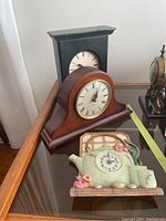 Photo shows three clocks including two wooden mantel clocks of different styles and a ceramic kitchen wall clock shaped like a teapot with flowers.