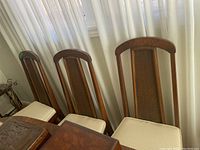 Set of six wood framed dining chairs with woven rattan back panels and cream upholstered seats, arranged in a row next to a curtain.