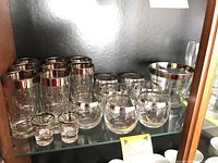 Image of glassware set with gold overlay including tall glasses, cocktail glasses, shot glasses, and ice bucket on a glass shelf.