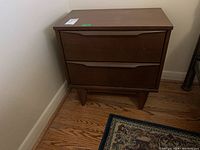 Wooden nightstand with two drawers closed, showing medium brown finish and angled legs on hardwood floor near wall.