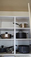 White shelves containing five pots: stainless steel saucepan, stainless steel casserole pot, black non-stick cast iron pot, a ceramic or cast iron pot, and a large grey non-stick covered pot.