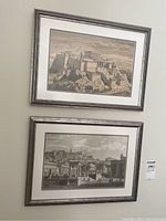 Two framed prints hanging on wall, shows the Acropolis and an ancient cityscape, label visible on side.
