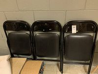 Three black metal folding chairs folded and standing against a wall with black vinyl padded seats and backrests.
