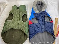 Top view of two dog coats side by side; olive green knit and quilted coat with zipper, blue puffy coat with faux fur hood.