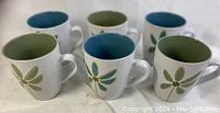 Set of six ceramic mugs arranged in two rows, three green and three blue, showing their flower design and interior color.