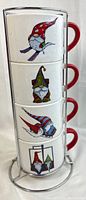 Front view of four white ceramic mugs stacked vertically on a silver metal rack. Each mug has a red handle and a colorful gnome illustration on the side.