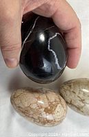 Hand holding black polished stone egg with white veining, next to two beige polished jasper-like stone eggs.