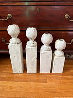 Four antique-style wooden candle holders of descending heights, painted white with distressed finish, placed on floor in front of wooden furniture.