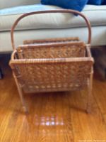 Front angle view of vintage rattan magazine rack showing woven design, curved handle, and four legs with slight unraveling on two legs.