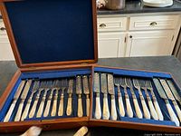 Full set of 12 forks and 12 knives with an extra larger knife in open wooden box lined with blue velvet.