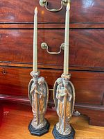 Pair of gold-tone angel candle holders holding tall taper candles, placed in front of a wooden chest of drawers with brass handles.