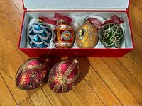 Top view of 4 Joan Rivers glass egg ornaments in box and 2 additional red gold filigree egg-shaped ornaments on floor