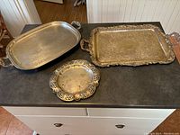 Three silver plated trays shown on grey countertop: large oval footed tray, smaller round pierced rim tray, and large rectangular engraved tray with decorative handles.