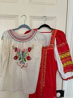 Two-piece lot showing the embroidered white blouse and red folk dress with colorful detailing hung on hangers