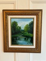 Full view of the vintage oil painting in the gold frame hanging on a white door, showing the landscape scene with river and trees and artist signature.