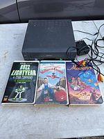 Front view of the black Goldstar VCR VHS player with power adapter, and three cartoon VHS tapes Buzz Lightyear of Star Command, Wallace & Gromit, and Aladdin lined up below it.