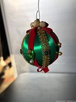 View of round handmade Christmas ornament wrapped with green satin ribbon, decorated with red velvet ribbon, gold scalloped trim, faux pearls, and green rhinestones, hanging by wire loop.