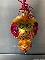 Front view of the glass Christmas ornament showing reflective hot pink circles and green bow detail on gold base.