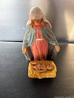 Mary figurine standing, wearing a pink dress and blue shawl with a white veil, facing downward.