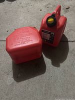 Two red plastic gas cans on concrete ground, one positioned upright showing yellow cap, the other laid flat showing label on bottom.
