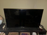 Front view of the 32 inch RCA flat screen TV turned off, placed on a black stand with a globe on the left and a globe-shaped light on the right.