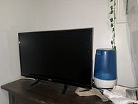 Black RCA 32 inch HD flat screen TV powered off, standing on a wooden surface next to a blue and white humidifier
