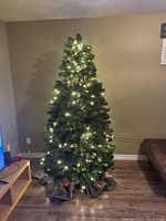 Photo showing a 6 foot artificial Christmas tree lit with warm white lights in a corner of a room near a TV and couch.