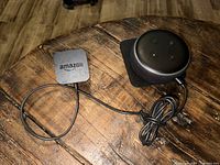 Top view of Amazon Echo Dot smart speaker and power adapter on wooden surface, showing control buttons and USB cable.
