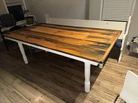 Full view of the rustic harvest table showing the wood plank top with black distressed accents and white turned legs on a wooden floor.