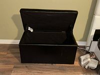 Black storage bench with open lid showing empty storage compartment inside against beige wall and wood floor.