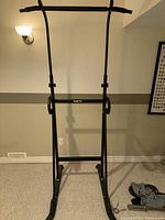 Black metal exercise station with pull-up bar and dip bars, set against a beige wall with a wall lamp and framed chart visible.