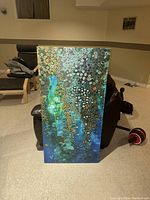 Photo of a large abstract painting placed upright on a leather armchair in a room, showing detailed multi-colored circular patterns on a blue-green background.