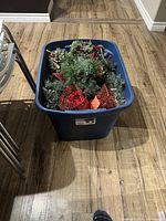 Blue plastic bin filled with assorted artificial Christmas decorations on wooden floor.