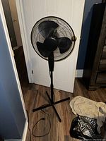 Photo of a black stand up oscillating fan with three blades, protective grill, height adjustable stand, and electrical cord on wooden floor against white door.