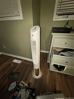 Photo of an upright white tower fan with a circular base standing on a wooden floor in a room corner.
