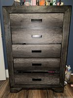 Front view of tall upright dresser showing five closed drawers with black handles and gray wood textured finish.