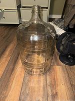 Large vintage glass carboy on wooden floor, showing body, neck, and embossed pattern.
