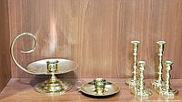 Entire lot of six brass candle holders shown on wooden shelf, including large chamber type with scroll handle, smaller chamber candle holder, and four traditional candlesticks.